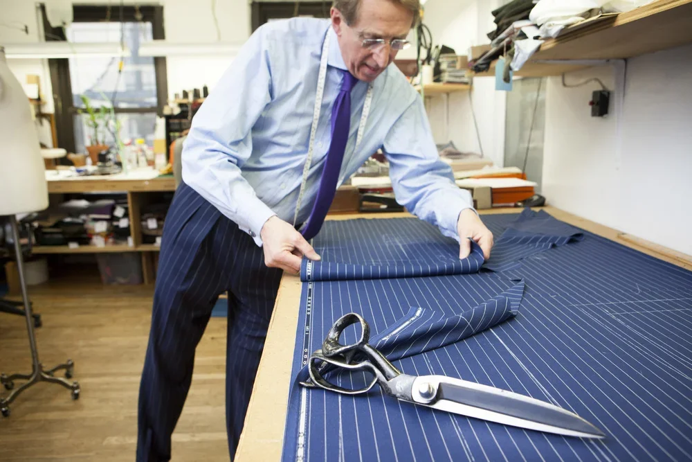 A tailor bent over the cutting board with heavy shears, marking a length of striped suiting.