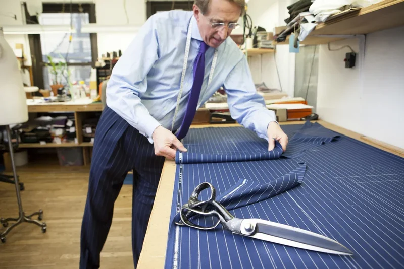A tailor bent over the cutting board with heavy shears, marking a length of striped suiting.